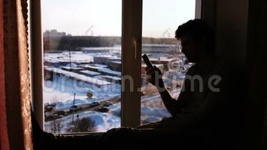 一个拿着手机坐在窗台上的男人的剪影。